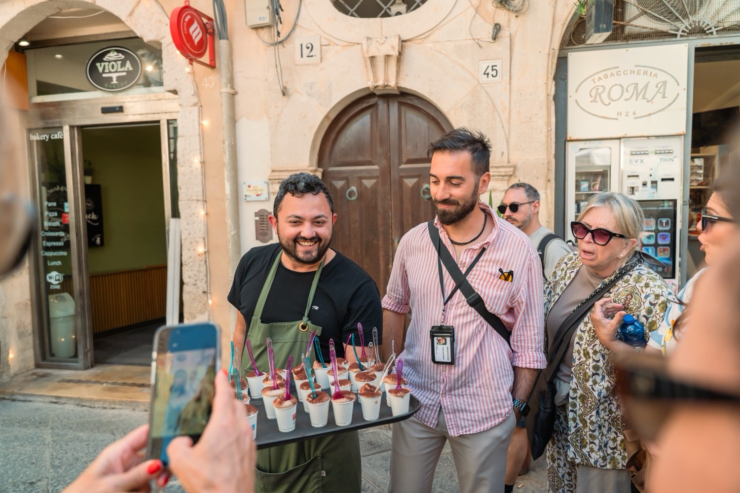 A Sicilian barista in an apron presenting a tray of traditional coffee to guests outside a historic bar in Ortigia
