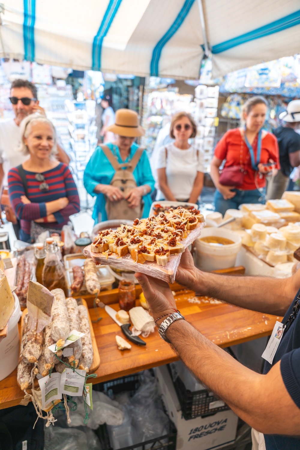 Hands offering a tray of capuliato bruschetta across a market counter laid with Sicilian cheeses and salami in Ortigia
